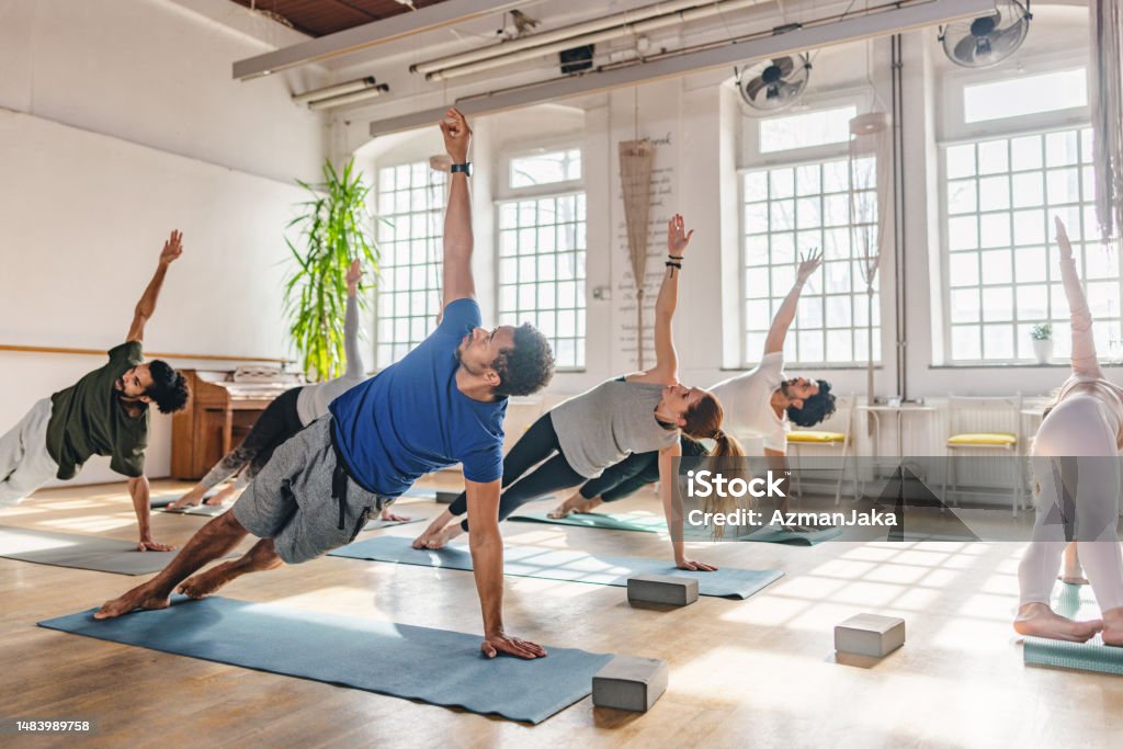 Diverse yoga practitioners doing a side plank on yoga mats during a yoga class. They are located in a beautiful yoga studio with natural light, big windows and plants.